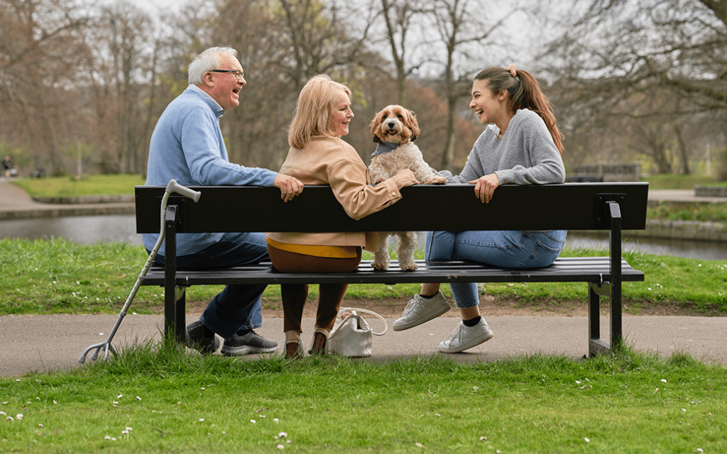Group of people sitting on a park bench laughing. A dog is sitting beside them. The group includes an older gentleman with a walking stick beside him.
